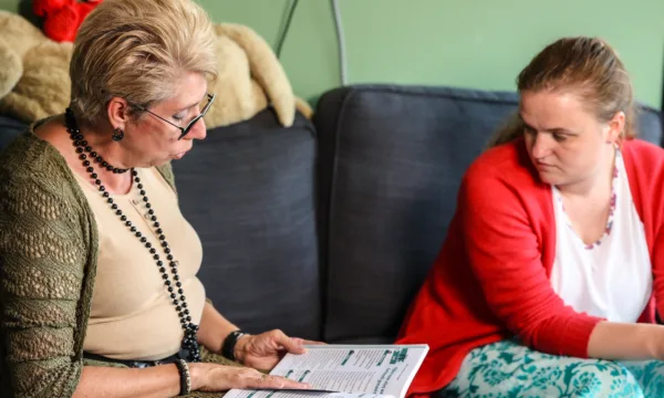 Twee mensen zitten op een bank. Eén persoon houdt een open boek of tijdschrift vast en beiden lijken in gesprek te zijn