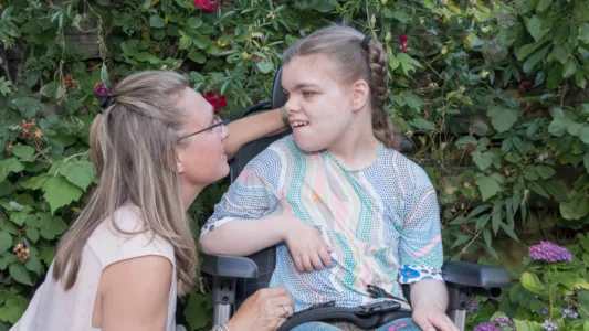 Een persoon in een rolstoel zit in een tuin met bloemen en struiken, terwijl een begeleidende persoon naast de rolstoel staat en iets tegen de persoon zegt.