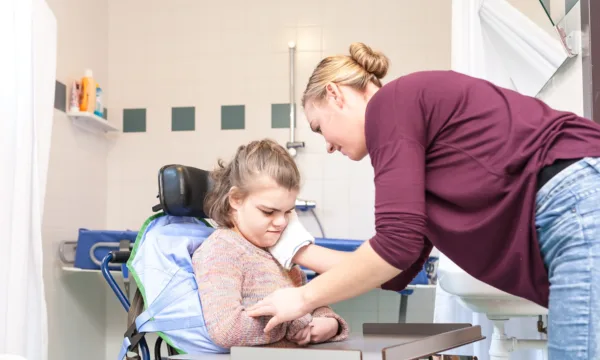 Een persoon ondersteunt een andere persoon in een rolstoel bij de wastafel in een badkamer, waar hulpmiddelen en verzorgingsmaterialen zichtbaar zijn.